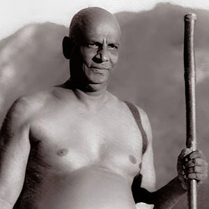 Sivananda standing by Ganges holding his water pot in one hand and his walking stick in the other. 
