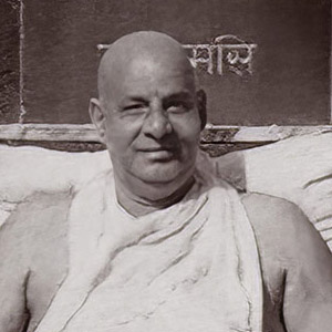 Sivananda sitting in front of the Sivananda Pillar upon which is written Serve, Love, Meditate, Realize. 