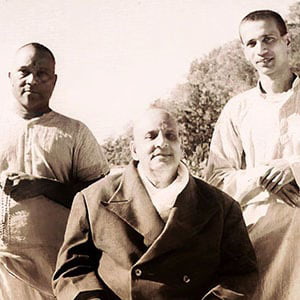 Swami Paramananda and Swami Chidananda standing behind Swami Sivananda (seated)